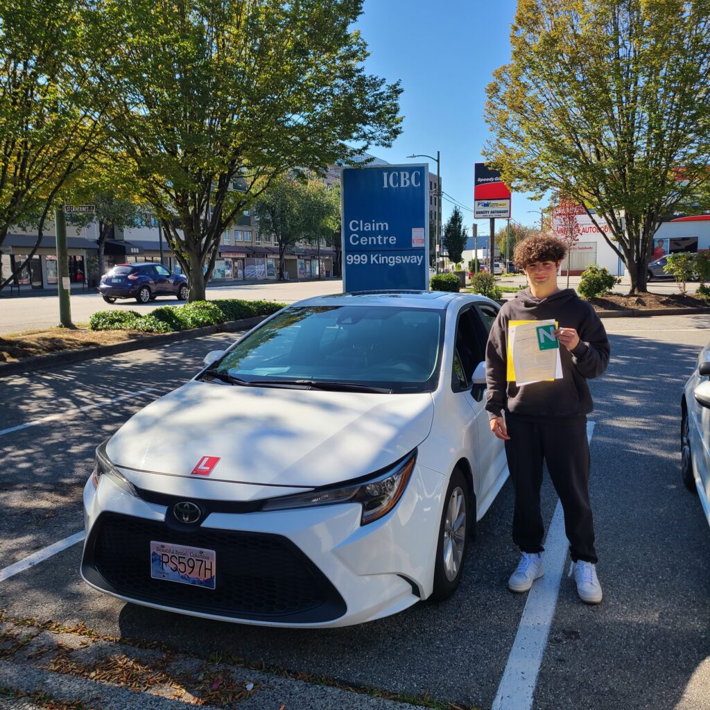 A man wearing a black hoodie with black joggers and white shoes holding papers standing beside white toyota corolla.